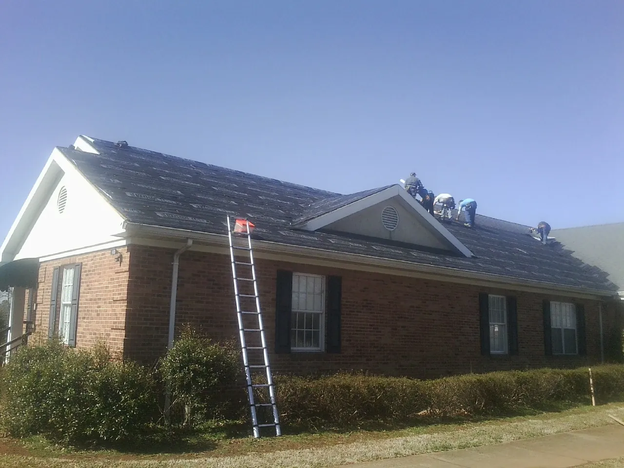 Crew tearing shingles off a brick ranch roof with a ladder set — Emergency Roof Repair work by our crew in Central Village
