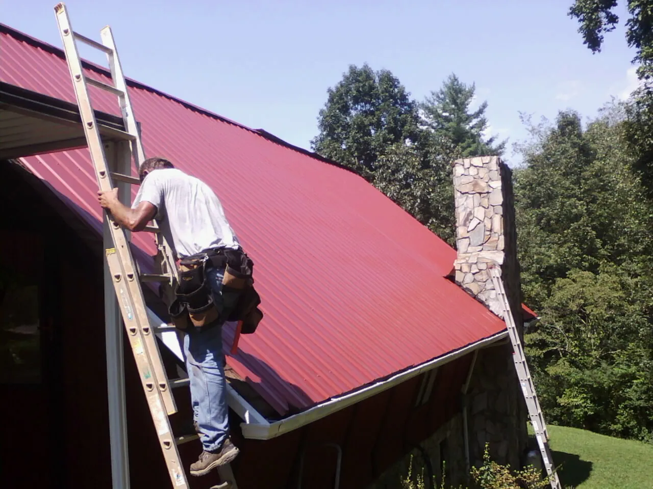Roofer on a ladder beside a red metal roof and stone chimney — Soffit & Fascia Repair by Roofing Brothers in Central Village